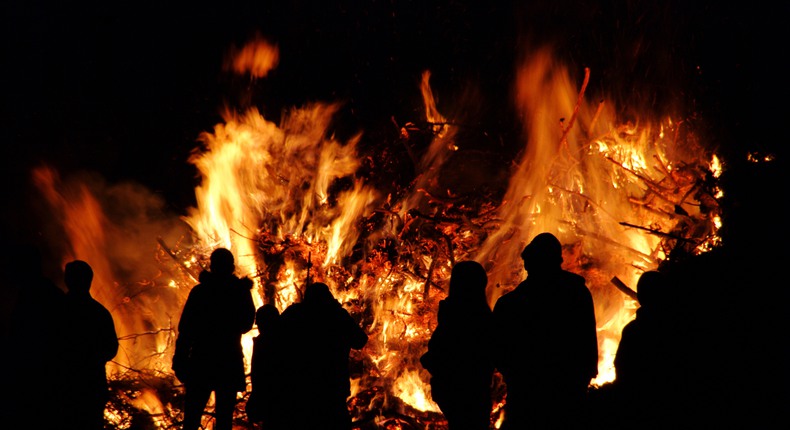 En stor brasa brinner om kvällen. Framför den syns siluetter av personer som tittar på elden.