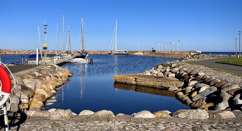 Blå himmel över Simrishamns hamn med segelbåtar och båtar förtöjda vid bryggorna. Livboj och stege i förgrunden.
