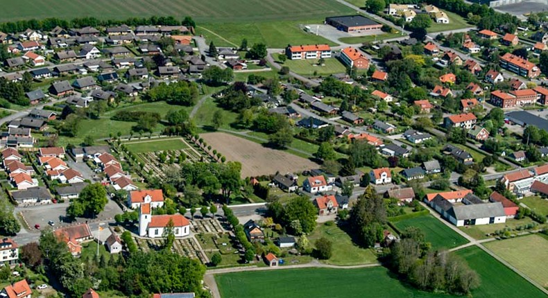 Flygfoto över St Olof med villor, verksamhetsbyggnader, kyrka, kyrkogård, träd, slingrande vägar - omgivna av fält och åkermark.
