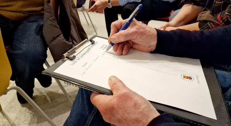 En man antecknar syna synpunkter på ett papper där det står "Trafik- och parkering i Kivik 2022". Pappret har stöd med ett svart hårt underlägg. Enbart mannens händer syns. Han håller i en blå penna. I bakgrunden syns stolar och benen från fler deltagande från mötet.