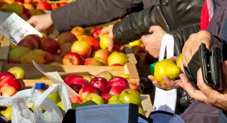 Ett handelsstånd med äpplen och päron i trälådor, På bilden syns händerna av tre kunder som plockar av frukten och en av dem håller i en svart plånbok för att betala.