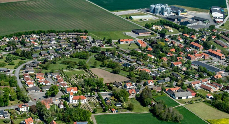 Flygfoto över St Olof med villor, verksamhetsbyggnader, kyrka, kyrkogård, träd, slingrande vägar - omgivna av fält och åkermark.