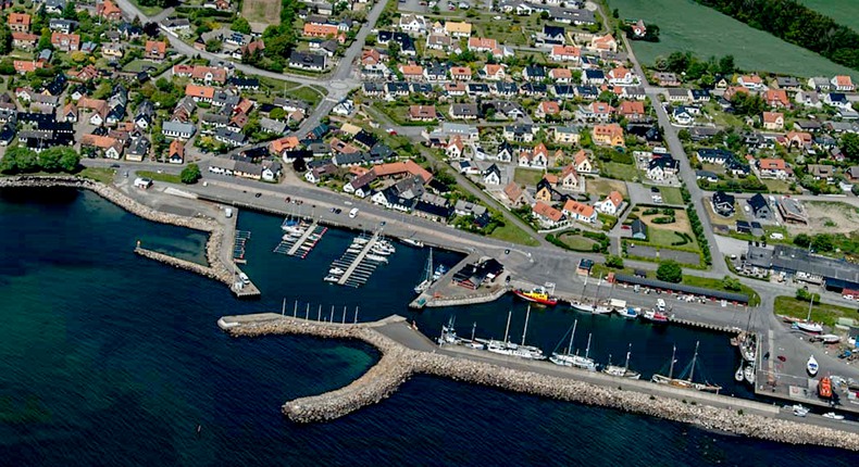 Flygfoto över Skilling by med hamn och båtar, slingrande gator, villor och verksamheter - omgivna av åkerfält.