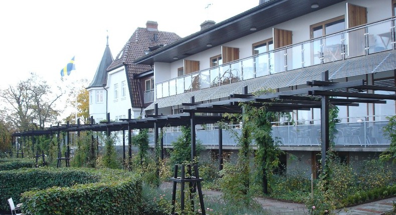 Bilden visar Kiviks hotell med inglasade balkonger i bakgrunden. I förgrunden en svart pergola som sträcker sig längs huskroppen. Framför den finns gröna buskar och planteringar.