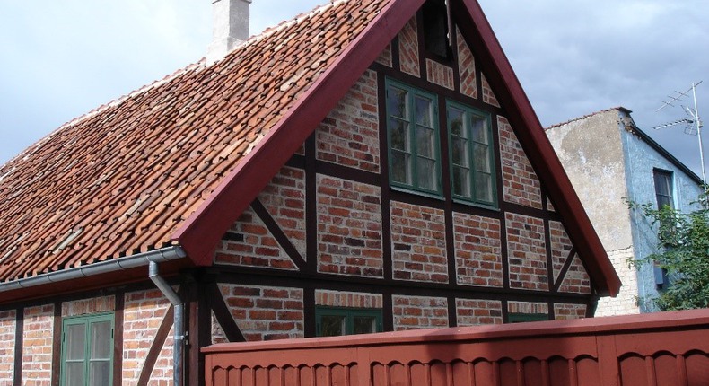 Del av hus sett snett från gavelsidan. Korsvirkeshus av tegel med tegeltak och gröna spröjsade fönster. På taket syns en skorsten och i förgrunden ett rött, högt, snidat staket.