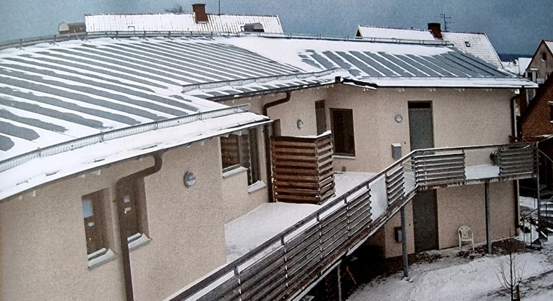 Foto av flerbostadshuset snödroppen i Simrishamn, taget snett ovanifrån. Två våningar. På husets grå plåttak ligger snö. På andra våningen finns loftgångar med räcke av trä. På innergården ligger ett tunt lager snö.
