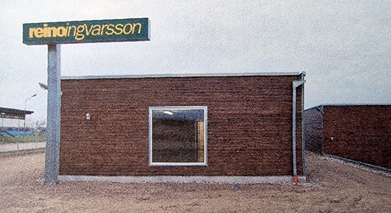En rektangulär träbyggnad med platt tak. Ett fyrkantigt fönster med vit karm är inbyggt mitt på fasaden. På husets vänstra sida står en skylt med företagsnamnet "Reino Ingvarsson". I förgrunden syns rödgul sand.