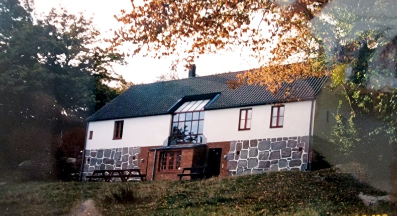 Putsat stenhus med svart tak och hög husgrund av stenar. Röda spröjsade fönster. Runt huset syns grönskande träd.