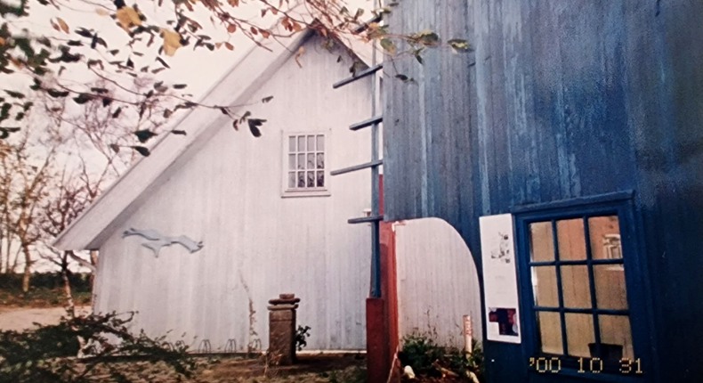 Två huskroppar av trä. Till vänster i bild syns en vit trägavel av en huskropp i 1,5 plan. På andra våningen visas ett fönster med vita, smala spröjs. Till höger i bild visas en hög träfasad som skiftar i blå toner. På bottenplan ett pröjsat fönster i samma kulör som huset.