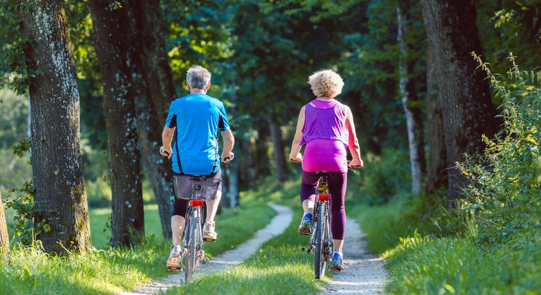 Två personer i grått hår och färgglada träningskläder cyklar på en skogsväg. 