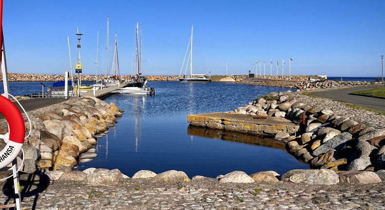 Blå himmel över Simrishamns hamn med segelbåtar och båtar förtöjda vid bryggorna. Livboj och stege i förgrunden.
