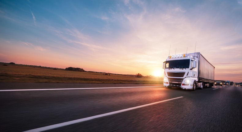 En vit lastbil kör på en landsväg i solnedgången. Bakom lastbilen syns personbilar som färdas i samma färdriktning.
