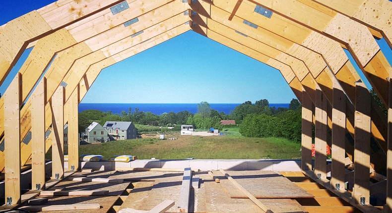 Ett hus håller på att byggas. Takstolarna i trä står bara i solen. Rakt fram syns en blå horisont.