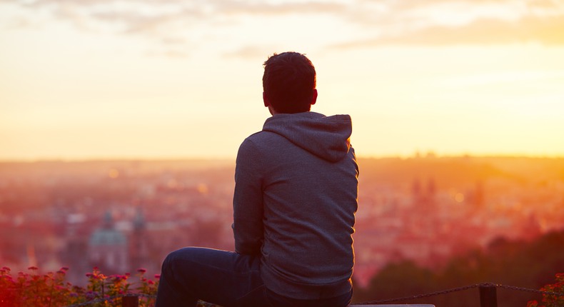En ung man sitter och tittar ut över en stad. Det är solnedgång. 