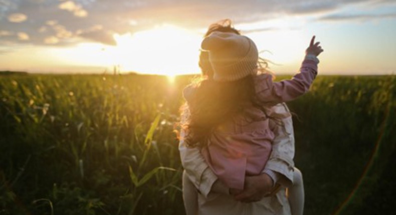 En liten flicka sitter i famnen på en förälder. Man ser ryggen på henne. Hon har mössa och rosa jacka. Det är solnedgång. 