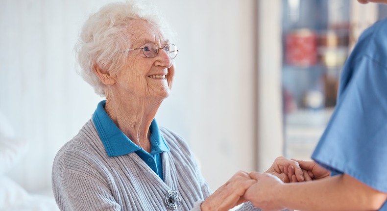 En äldre kvinna sitter på sin säng. Hon ler och håller en sköterska i händerna. 