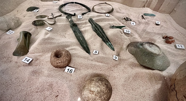 Forntidsverktyg som ser ut som flintayxor och metallknivar ligger upplysta på sand i glasmonter som är nedsänkt i ett golv.